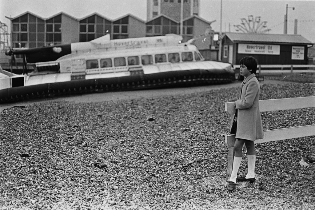 A young woman stands on a rocky beach looking out over the water. Behind her is a hovercraft parked on the beach